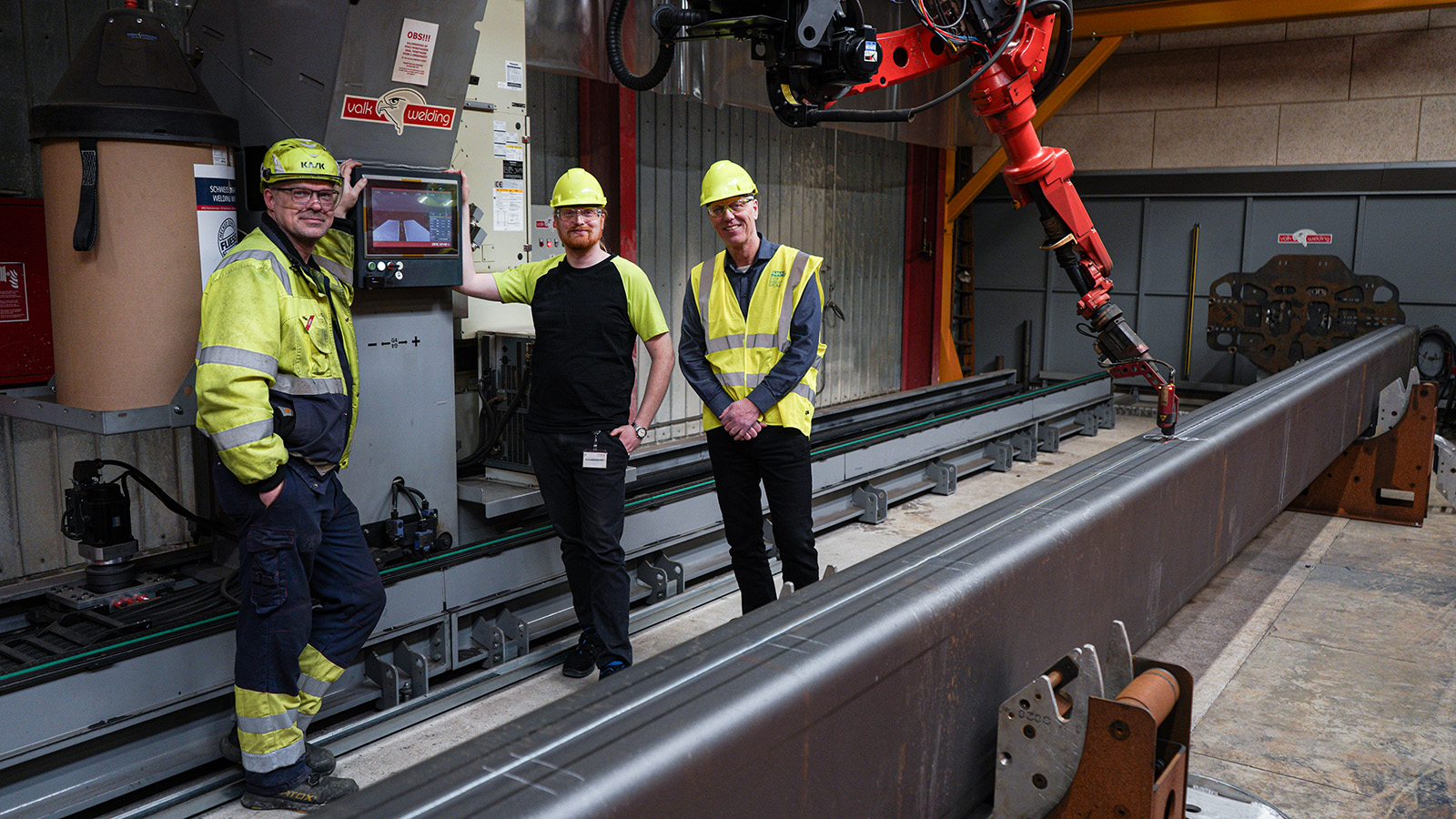 Three employees are standing in front of a screen in a robotic welding cell.